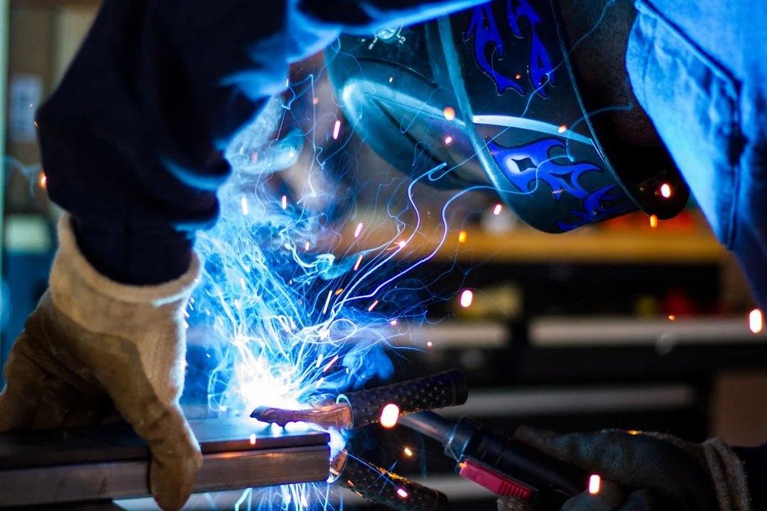 A professional welder in full PPE lays a clean root pass on a structural steel joint inside a fabrication shop, with a Miller multi-process machine visible in the background